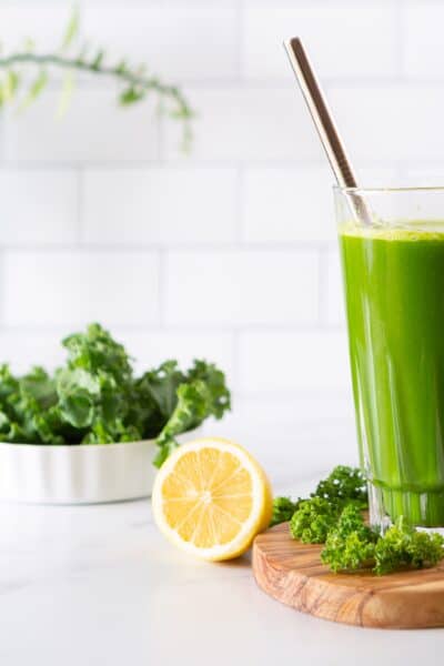 glass of green juice on a cutting board in a white kitchen with kale and lemons in the background