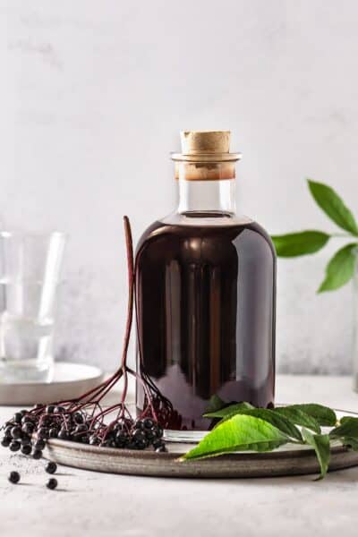 Front view of elderberry syrup in a glass bottle light gray background with fresh elderberry on tray.