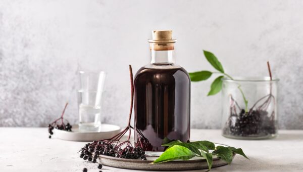 Front view of elderberry syrup in a glass bottle light gray background with fresh elderberry on tray.