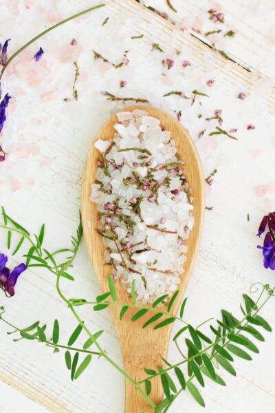 wooden spoon with salt chunky salt surrounded by herbs and purple flowers on a white wooden background