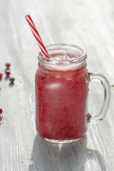 bowl of cranberries next to a cranberry smoothie in a jar with a red and white striped straw and herb leaves in the background