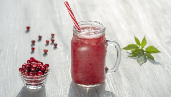 bowl of cranberries next to a cranberry smoothie in a jar with a red and white striped straw and herb leaves in the background