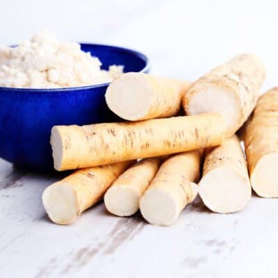 Clean and cut horseradish root next to a blue bowl of horseradish paste on a white wooden background