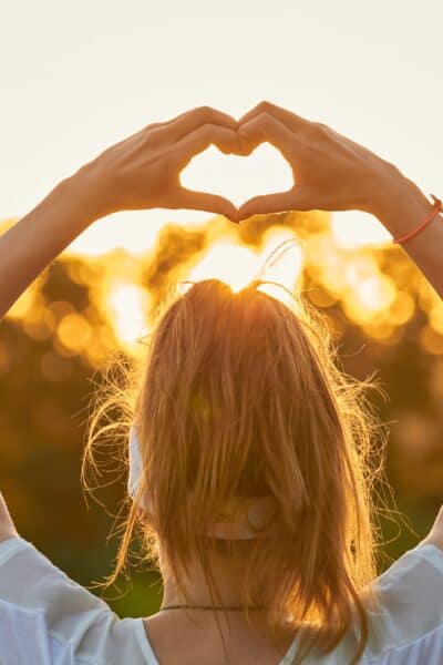 Woman in nature holding heart-shape symbol made with hands at sunset.